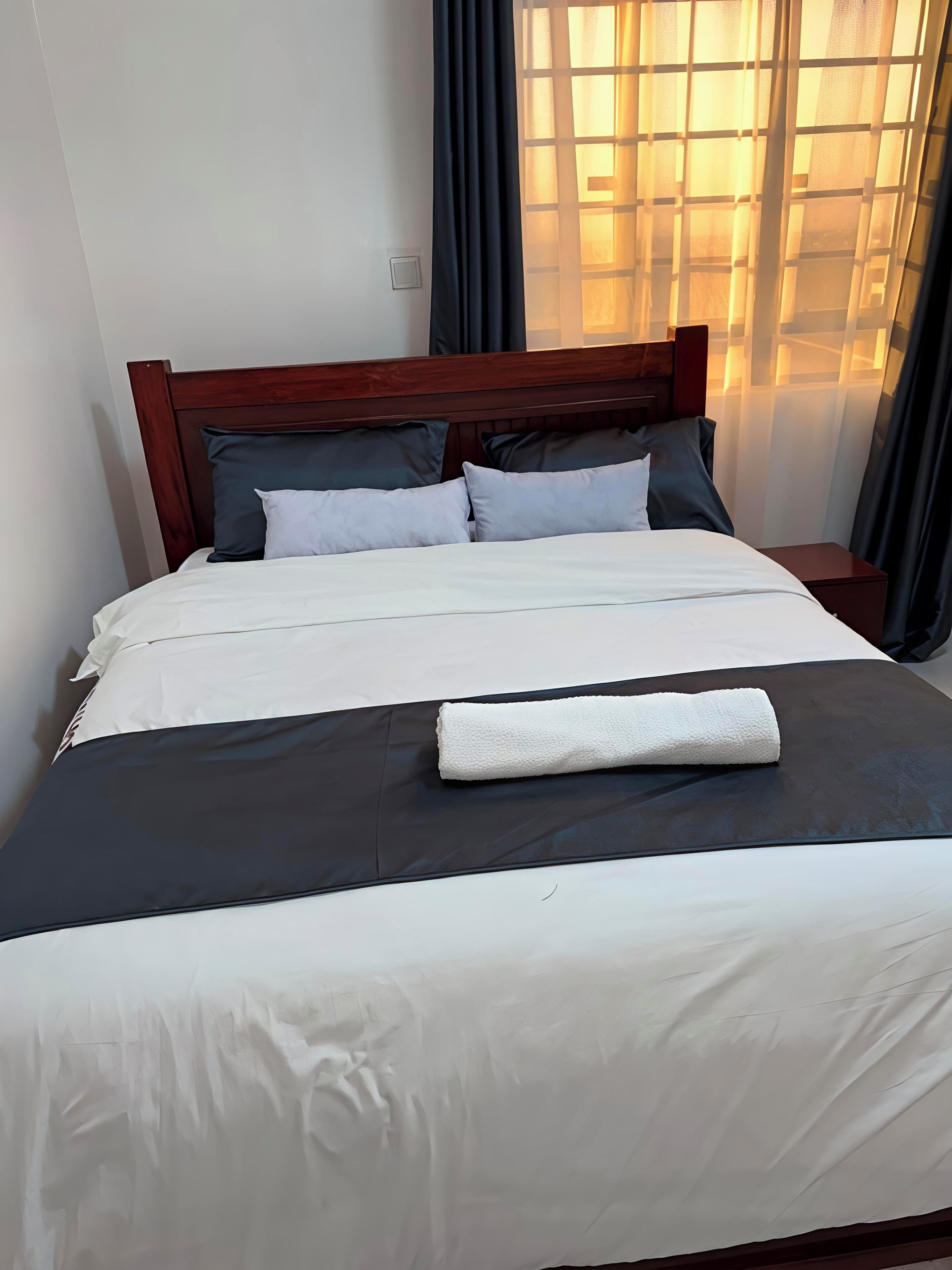 Una cama bien tendida con una colcha blanca y oscura, una almohada blanca y una toalla blanca doblada encima.