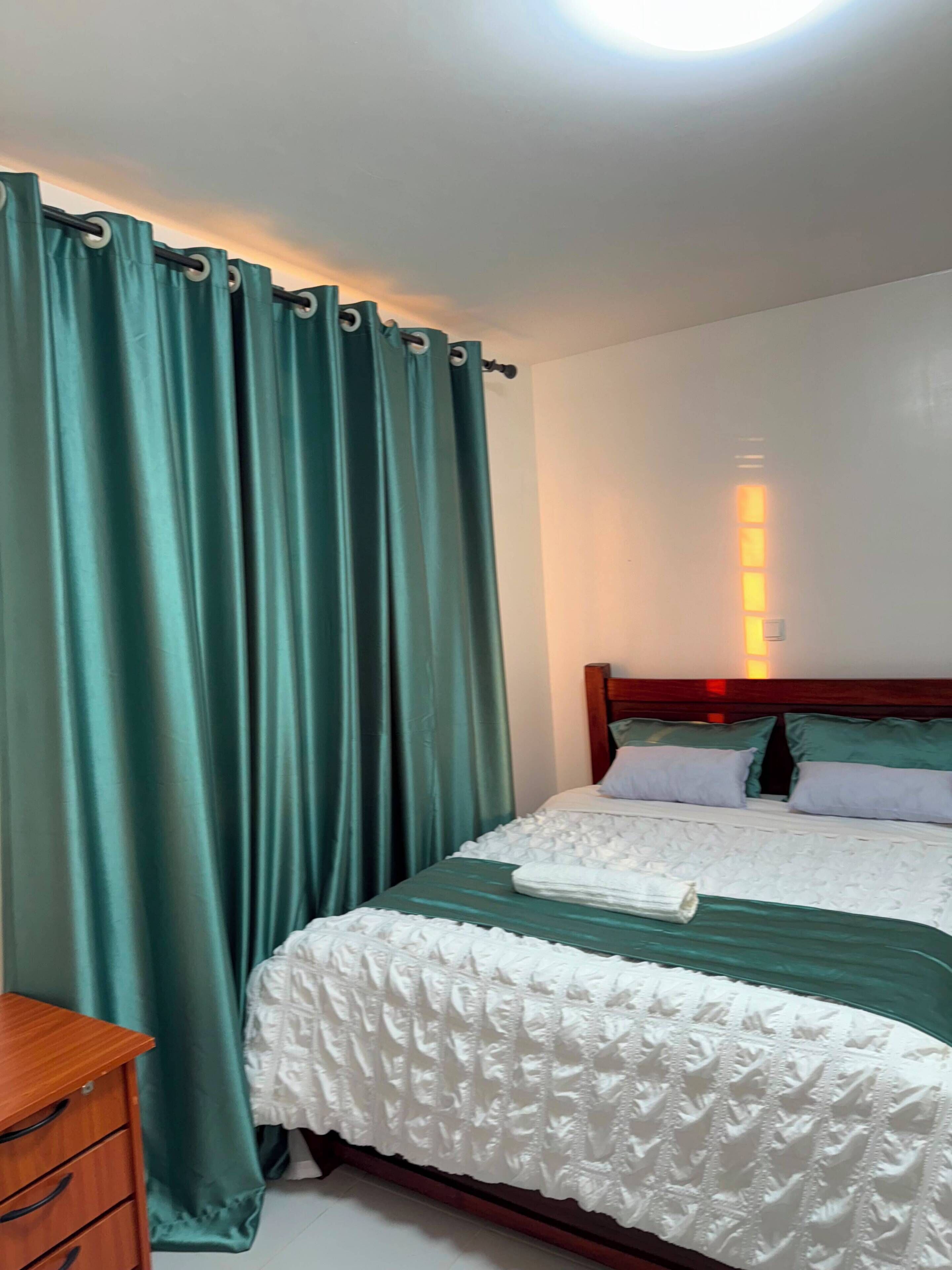 A hotel room with a bed, curtains, a wooden nightstand, and a wall-mounted light fixture.