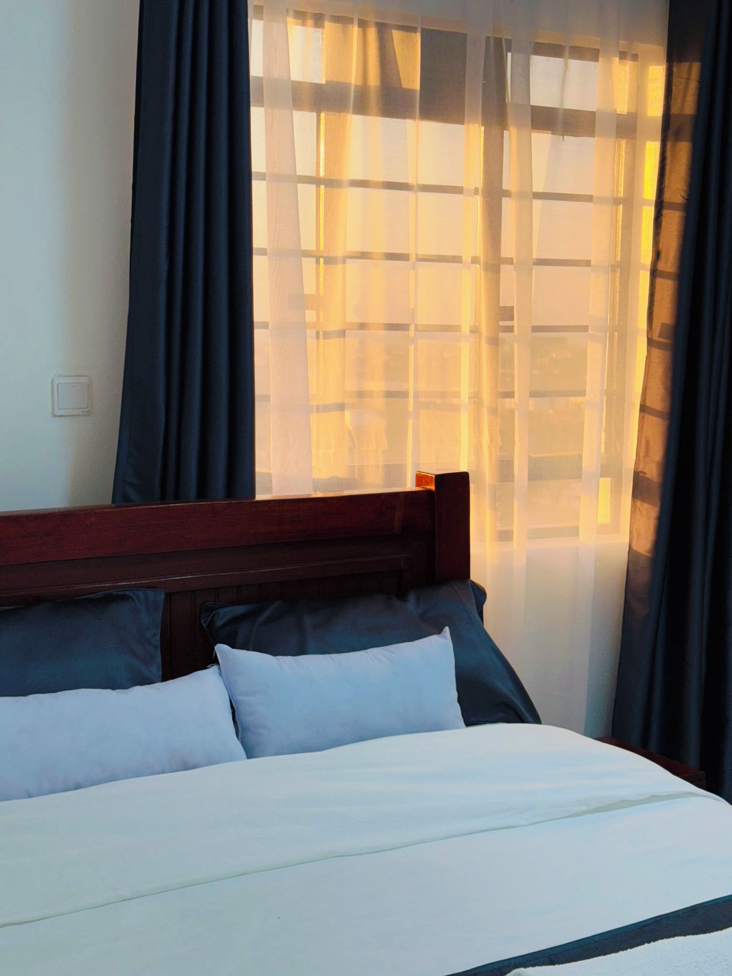 A bed with a wooden headboard, white bedding, and dark curtains partially drawn.