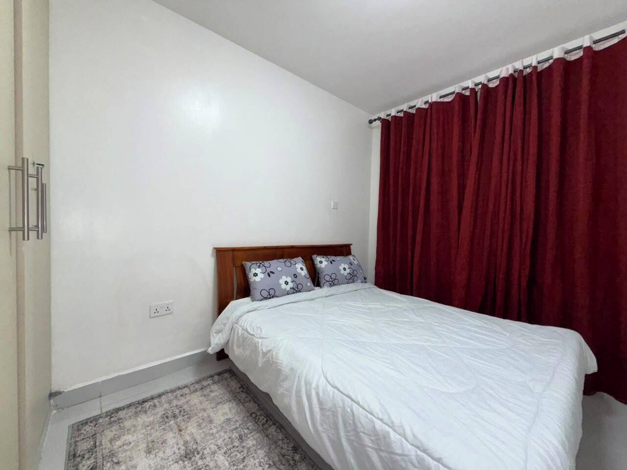 A bedroom with a single bed, a wooden headboard, two patterned pillows, a red curtain, and a white wall.