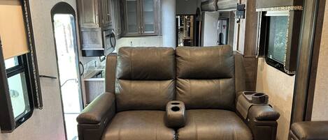 A compact living space with a leather sofa, a ceiling fan, and a kitchenette.