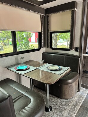 Ett kompakt inre utrymme i husbilen med en matplats som inkluderar ett bord och en bänk, stora fönster med utsikt över grönområden och ett litet köksutrymme.