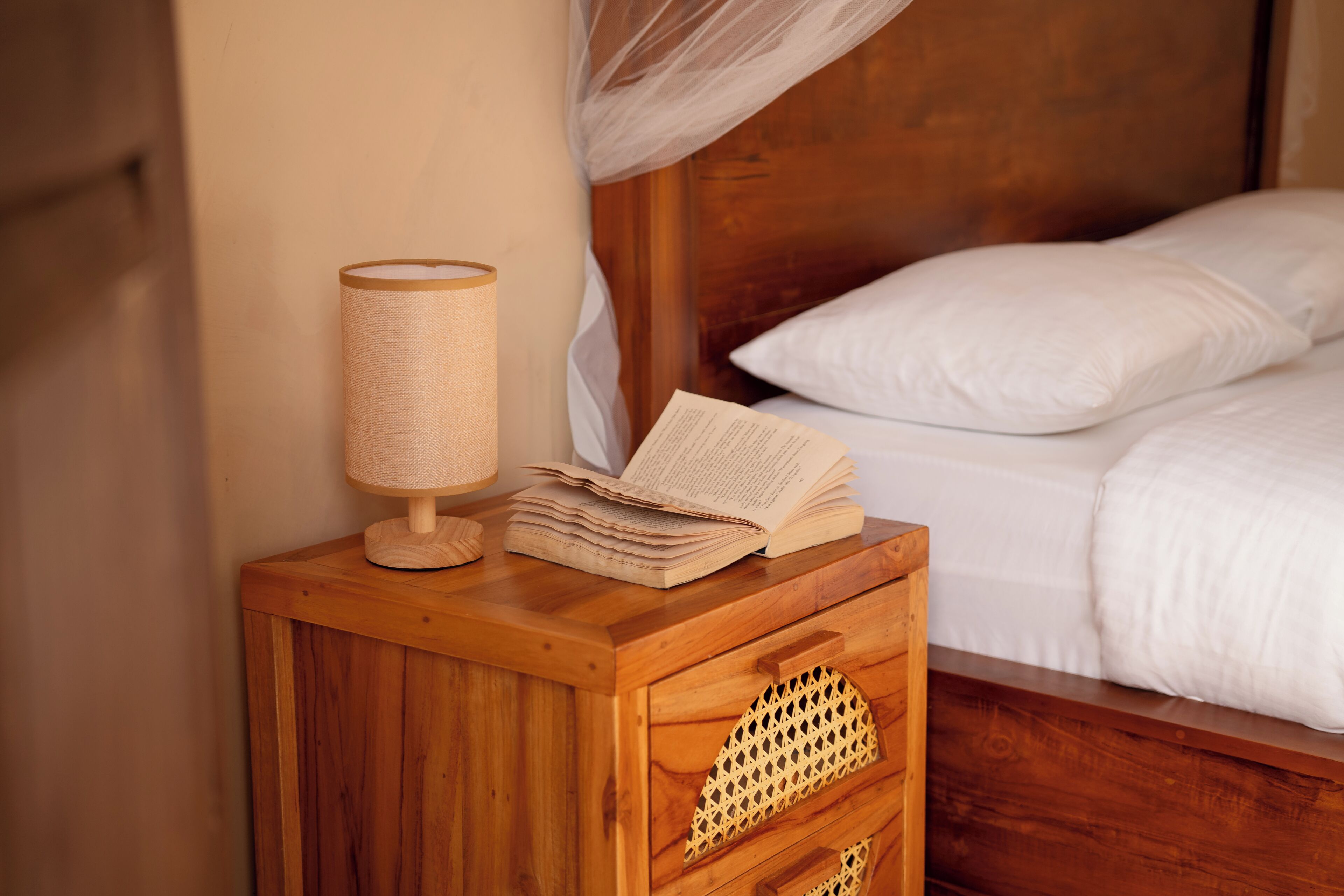 A wooden bedside table with a lamp, an open book, and a bed with white bedding.