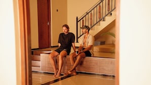 Two individuals seated on a wooden bench in a well-lit indoor space with a staircase and a cushioned seating area in the background.