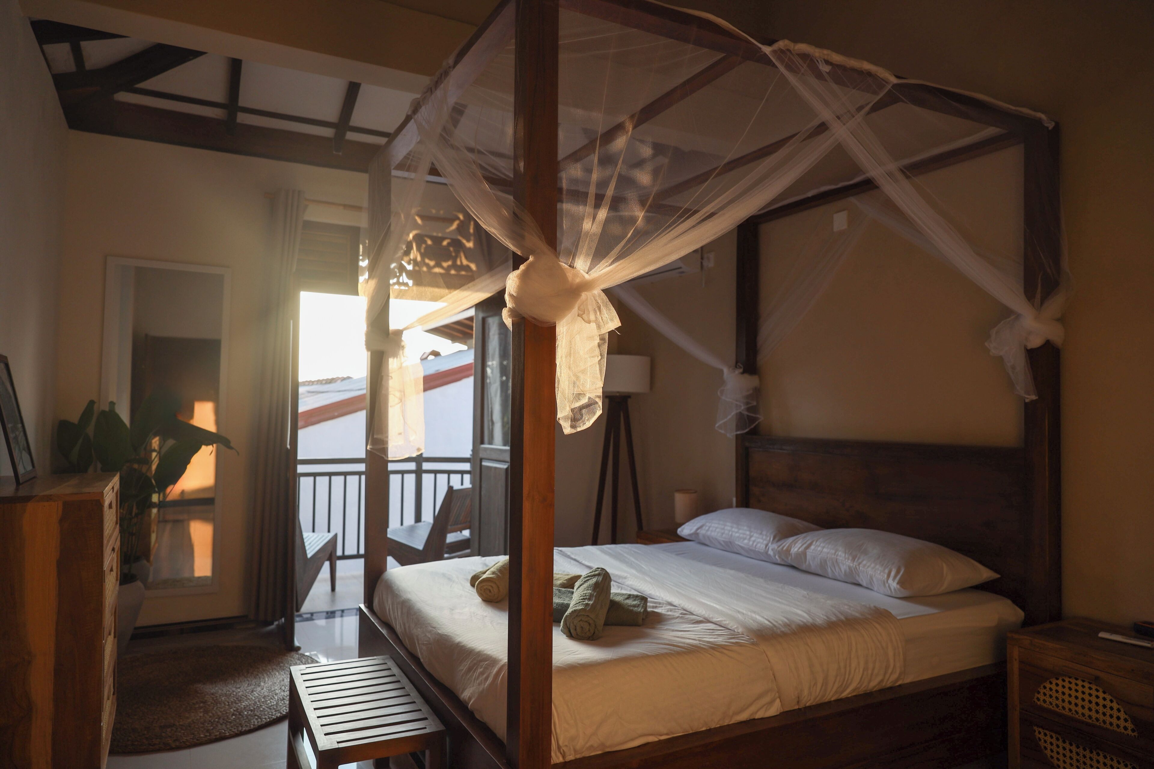 A four-poster bed with a canopy, a bedside table, a chair, and a view of the outside balcony.