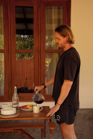 A person pouring coffee into a mug on a wooden table with a bottle and fruit bowls.