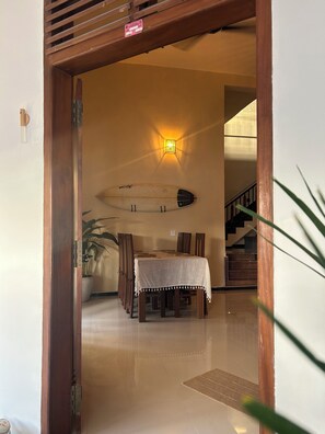 An interior view of a dining area with a table set for four, a wall-mounted surfboard, and a staircase in the background.