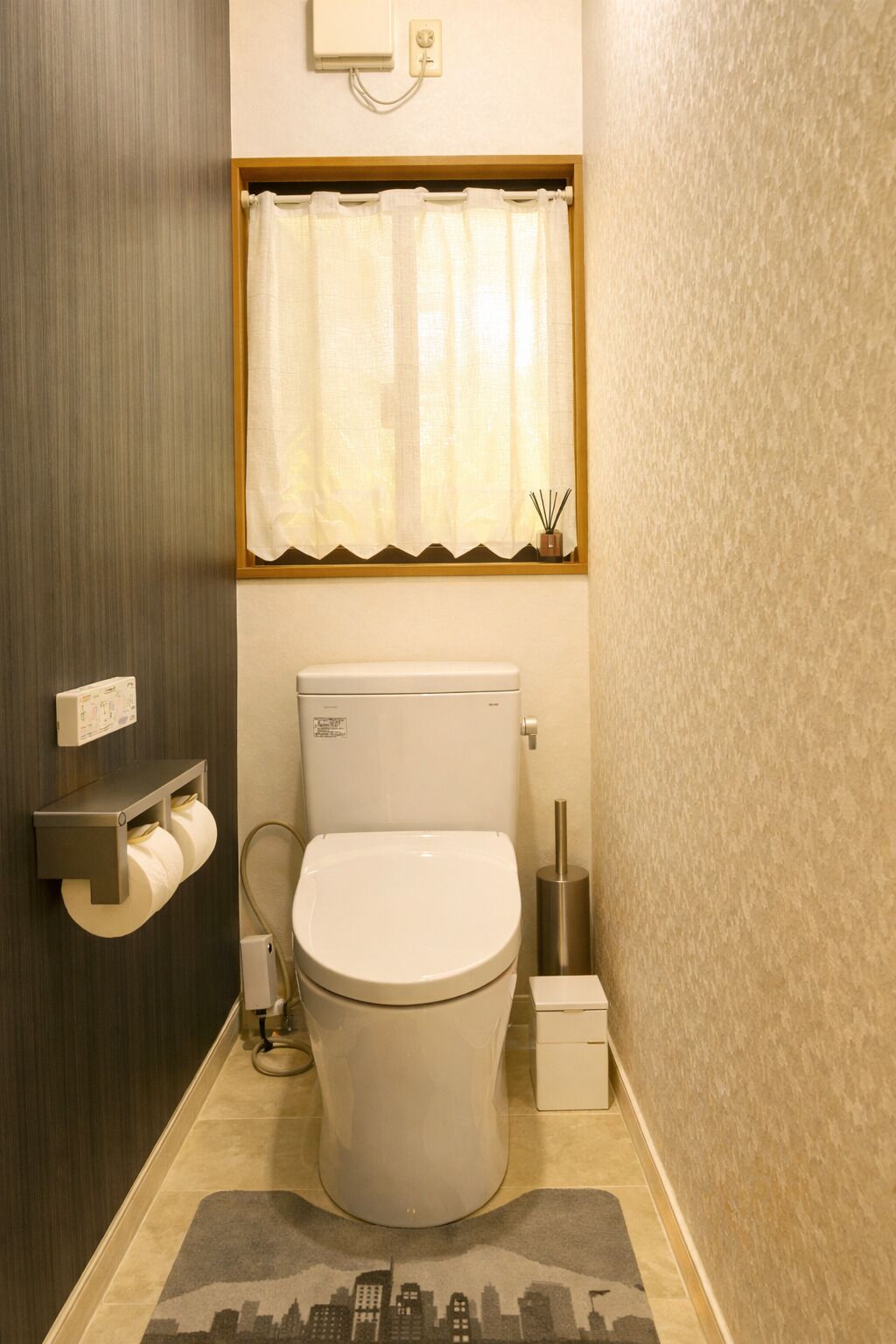A small bathroom with a toilet, a toilet paper holder, a window with a curtain, and a small potted plant.
