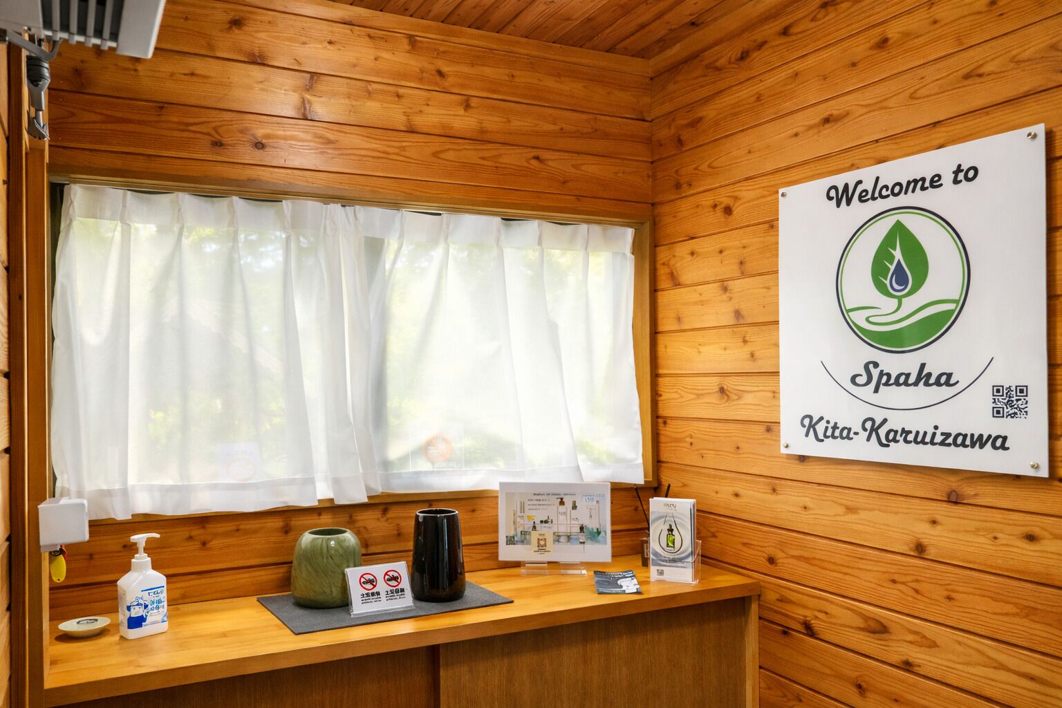 Interior de una cabaña de madera con una mesa de recepción, un letrero de bienvenida, una ventana con cortinas y un dispensador de gel antibacterial.
