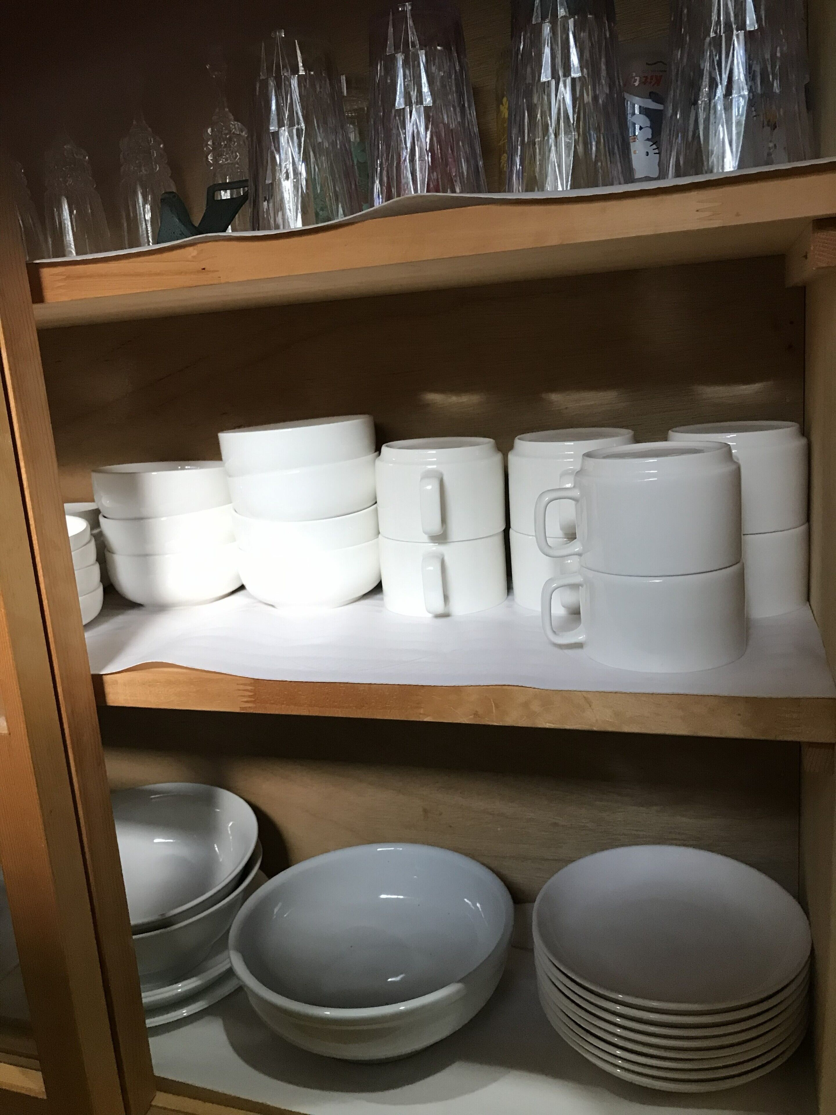 Une étagère avec des tasses et des bols en céramique blanche, et une étagère en bois au-dessus, sur laquelle sont disposées des bouteilles en verre transparent.