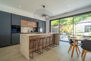 A modern kitchen with a central island, bar stools, and a dining area with a view of greenery. - Villa Les Patys (Vouvray)