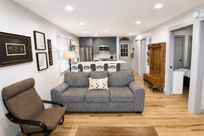 Un salon moderne avec un canapé gris, un fauteuil marron et un banc en bois. On aperçoit, au fond, une cuisine avec des meubles gris et des appareils électroménagers en acier inoxydable.