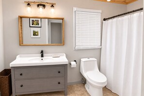 Une salle de bain avec un lavabo blanc, des toilettes, un miroir orné de photos et une fenêtre avec des stores.