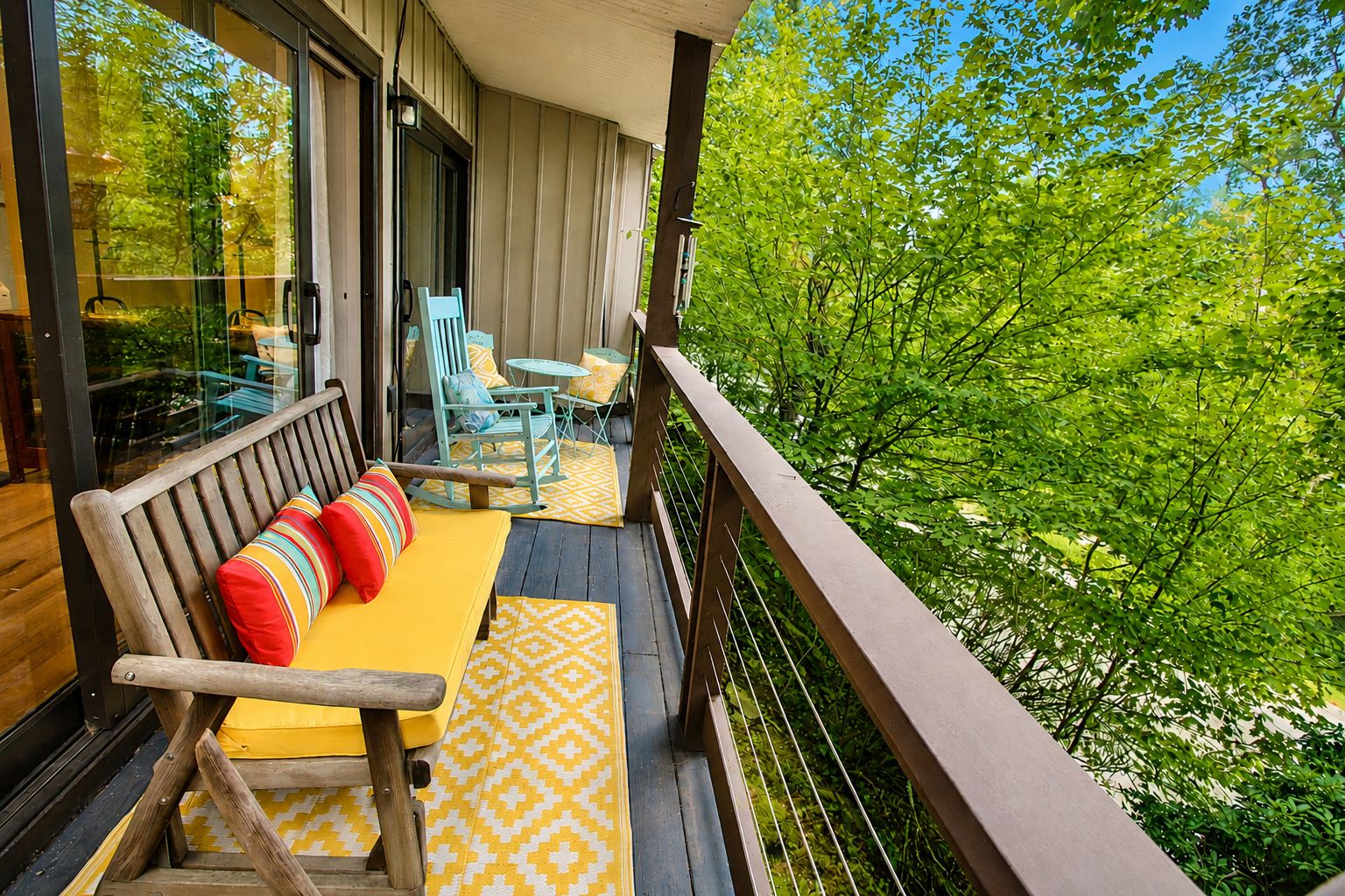A wooden deck with a bench and chairs, a glass door, and a view of trees.