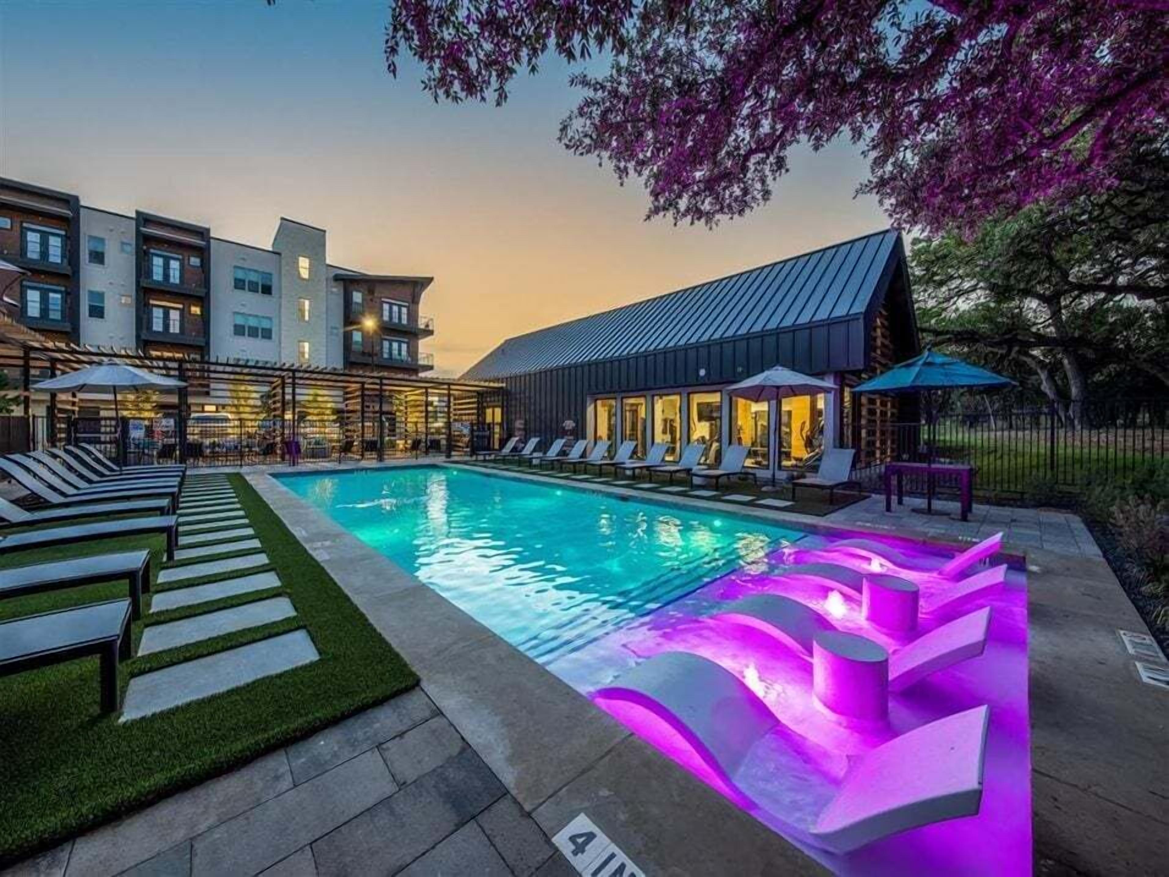 Una piscina con sillones de descanso y una zona de estar cubierta, rodeada de edificios de arquitectura moderna.