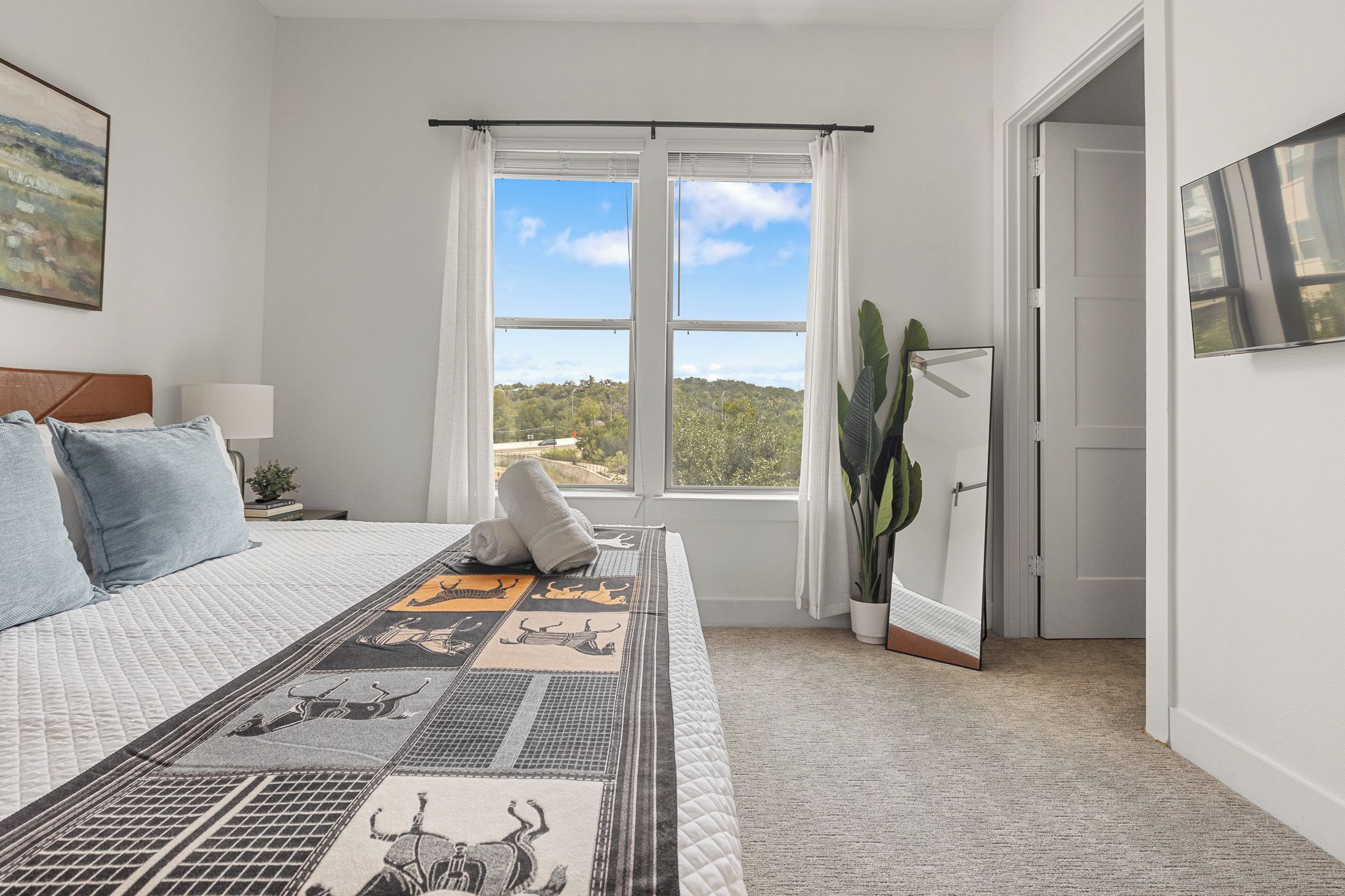 A bedroom with a large bed, a window with a view, a door, and a plant.