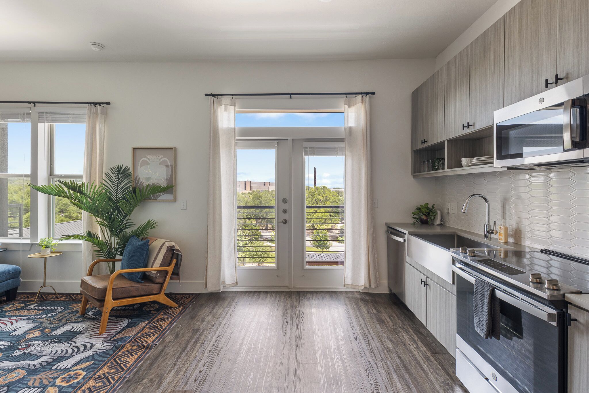 A modern kitchen with a built-in oven and microwave, a dining area with a blue sofa and a patterned rug, and a large sliding glass door leading to a balcony with a view of trees and buildings.