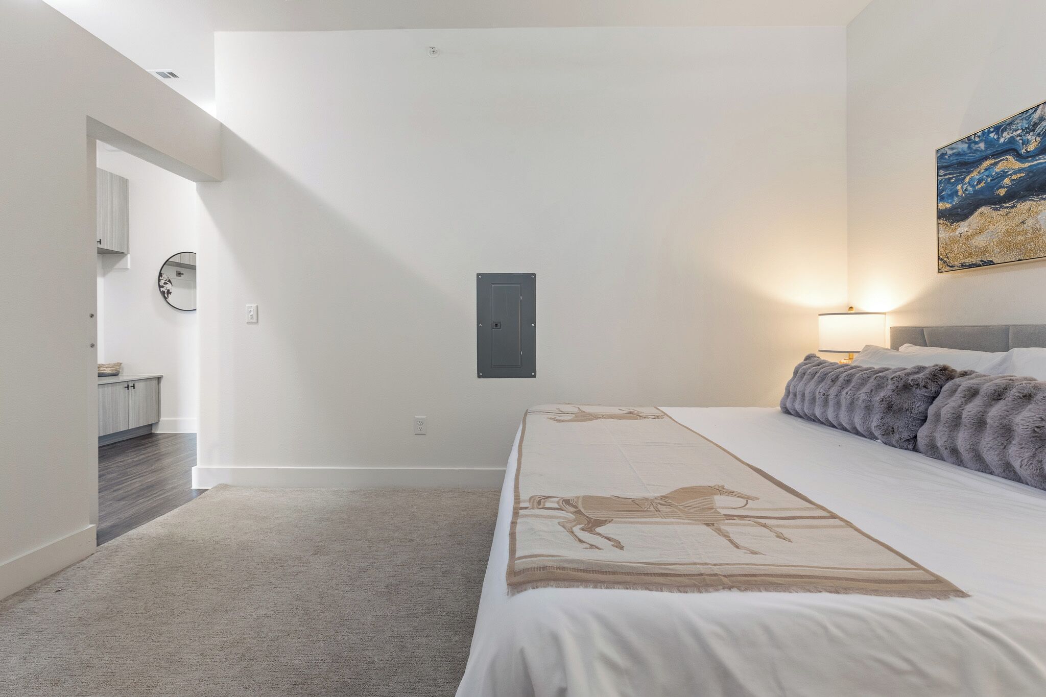 A bedroom with a bed, a nightstand, a lamp, a painting, and a door leading to another room.