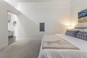 A bedroom with a bed, a nightstand, a lamp, a painting, and a door leading to another room.