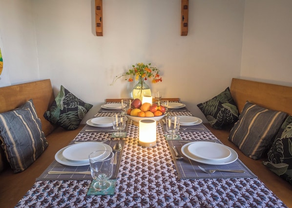 A dining table set with plates, glasses, and a fruit bowl, surrounded by patterned placemats and cushions.
