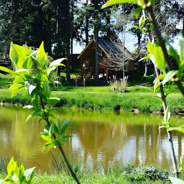 A wooden cabin with a staircase leading to the entrance, surrounded by lush greenery and a calm pond.