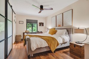A bedroom with a bed, bedside table, lamp, window, and artwork on the wall.