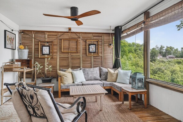 Une pièce chaleureuse avec un ventilateur de plafond en bois, un canapé avec des coussins, une petite table et une vue sur la verdure à l’extérieur.