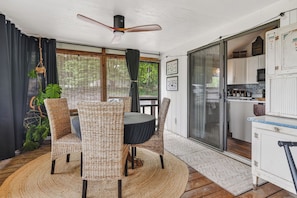 A room with a ceiling fan, wicker chairs, a round table, a view through a glass door to a kitchen, and a large plant.
