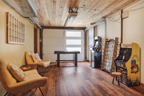 A room with wooden flooring, a pool table, two leather chairs, a foosball table, and a Pac-Man arcade machine.