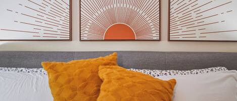 Ein Bett mit zwei Kissen, einem grauen Kopfteil und einer Wand mit drei gerahmten Kunstwerken.
