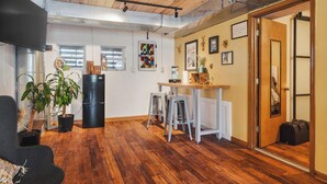 A room with wooden flooring, a black refrigerator, a dining table with stools, and a door with a suitcase.