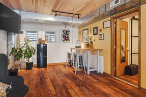 A room with wooden flooring, a black refrigerator, a dining table with stools, and a door with a suitcase.