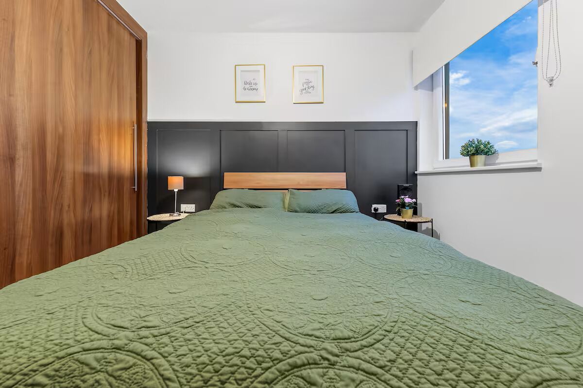 Un dormitorio con cama, mesita de noche, lámpara, ventana con planta y dos cuadros en la pared.