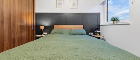 Un dormitorio con cama, mesita de noche, lámpara, ventana con planta y dos cuadros en la pared.