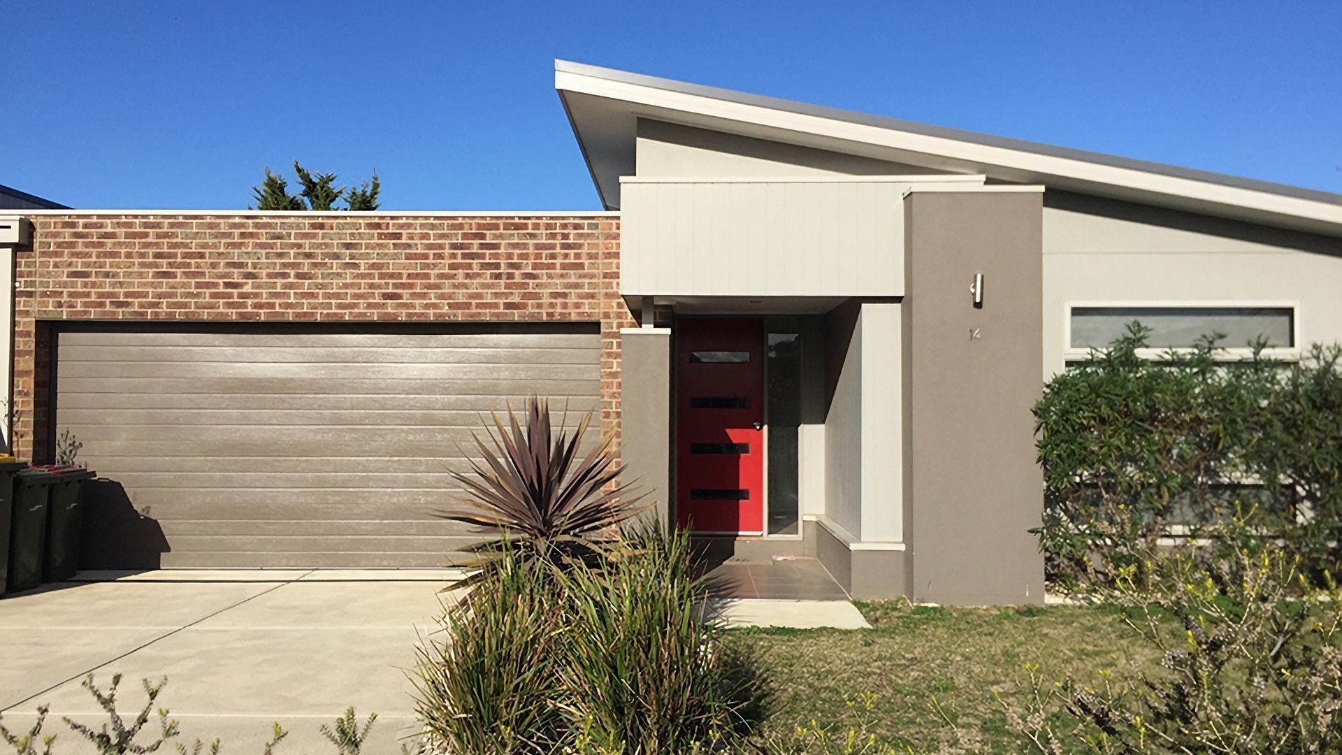 Une maison moderne à deux étages, dotée d’un garage, d’une porte rouge et d’une façade en briques.
