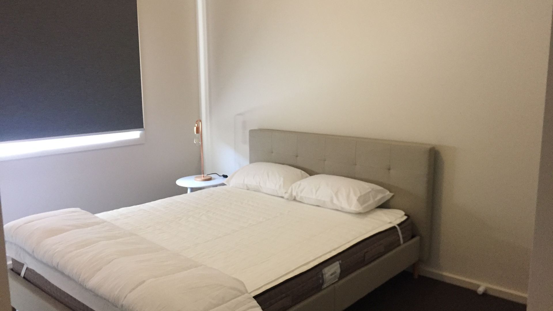 A neatly made bed with a grey headboard and white bedding in a room with a small bedside table and a lamp.