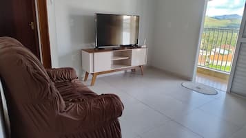 Uma sala de estar com um sofá marrom, um rack branco com uma televisão de tela plana e vista para uma paisagem verde através de uma porta de vidro.