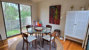 A dining area with a glass table surrounded by chairs, a colorful cushion, and a view through large windows. - Moonah Delight - Point Lonsdale (Point Lonsdale)