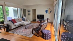 Ein großzügiges Wohnzimmer mit einer Couch, einem Sessel, einem Hocker, einem Fernseher und einem großen Fenster.