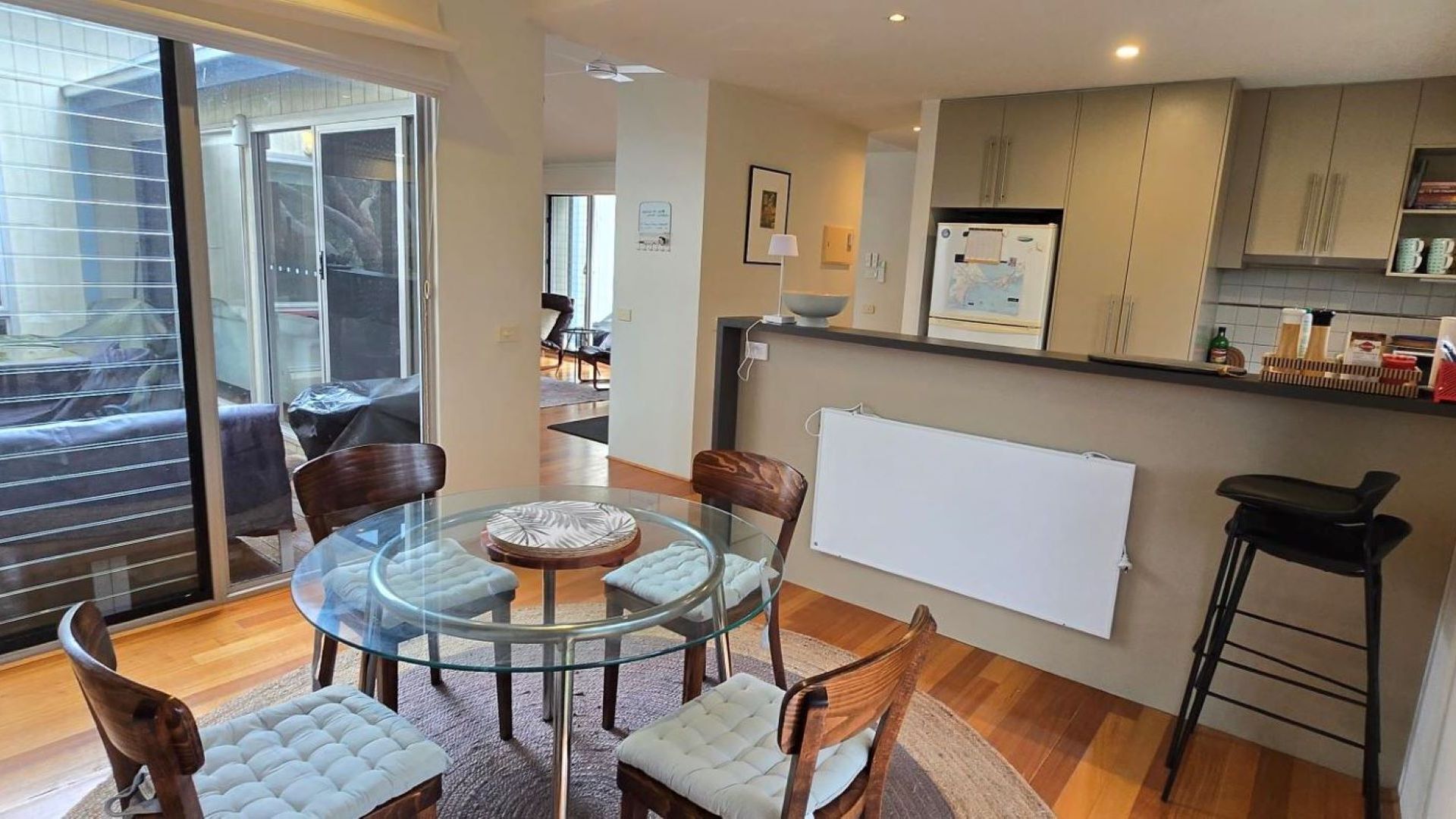A modern kitchen with a glass dining table, wooden chairs, and a refrigerator.