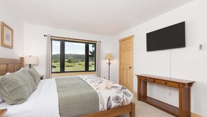 Quarto com cama, mesa de cabeceira, luminária, televisão e janela com vista para área verde.