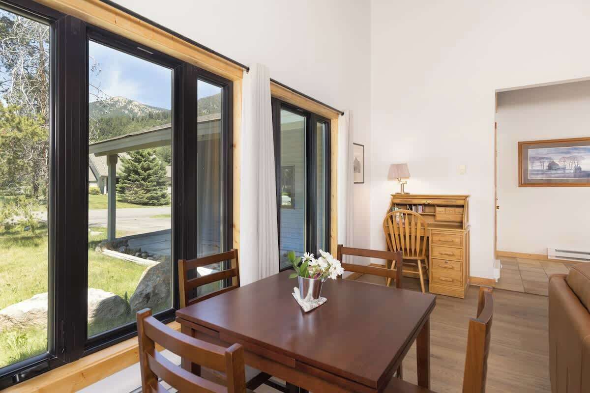Uma sala de jantar com mesa e cadeiras, uma escrivaninha de madeira com abajur, e vista para o exterior através de amplas janelas.