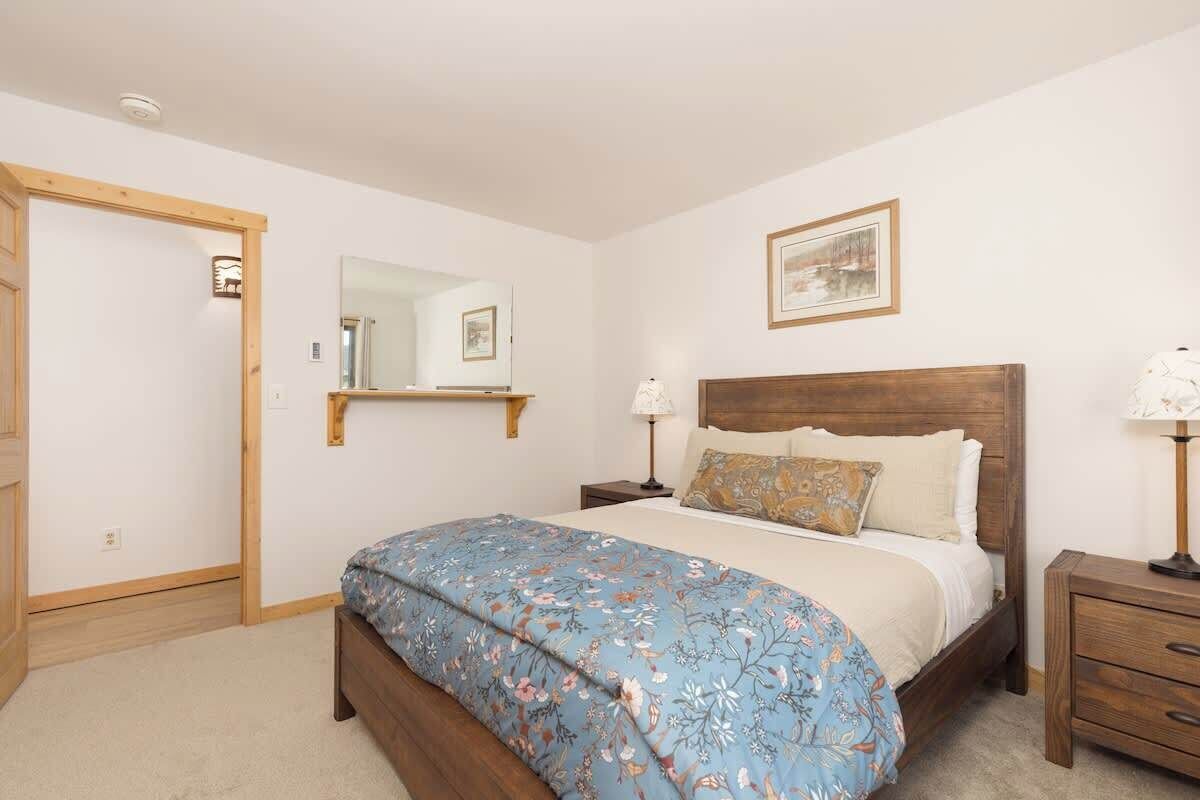 A bedroom with a wooden bed, a nightstand, a bedside table, a mirror, and a framed picture on the wall.