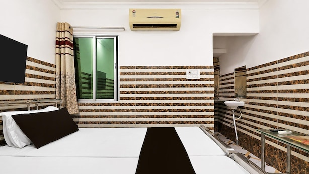 Habitación de hotel con una cama, ventana con cortinas, equipo de aire acondicionado, espejo y una pequeña mesa con lámpara.