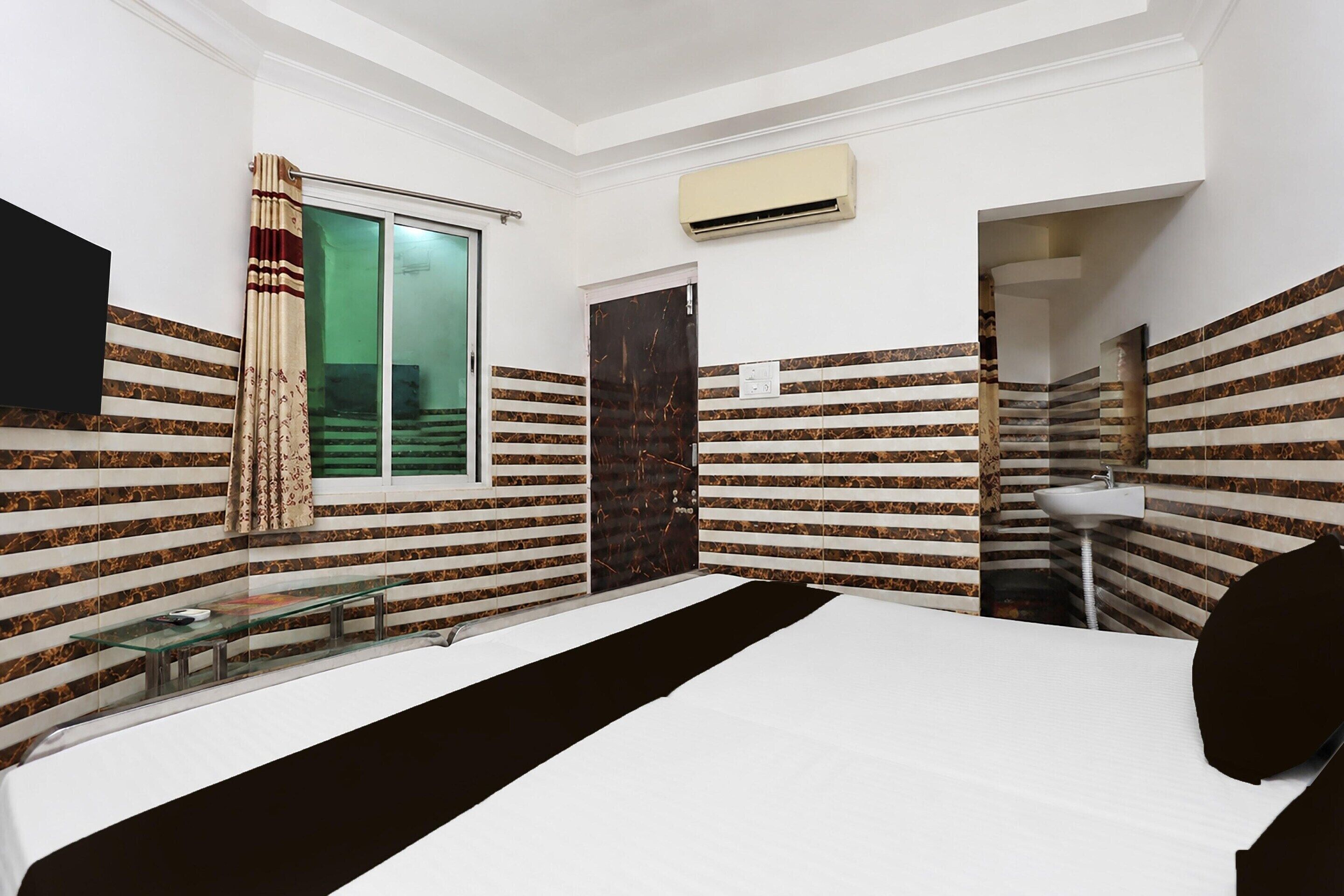 A bedroom with a bed, a window with curtains, an air conditioning unit, and a bathroom with a sink and mirror.