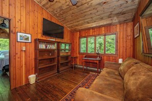 Ein Zimmer mit Holzwänden, einem Flachbildfernseher, Bücherregalen, einer Couch und großen Fenstern.