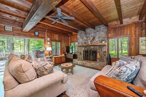 A log cabin interior with a stone fireplace, wooden ceiling beams, and large windows. - New! Lake Front Home w/ Private Dock! (Swanton)