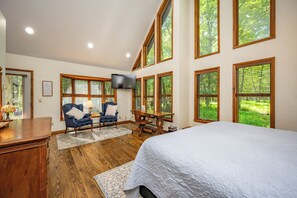 Une chambre dotée d’un grand lit, d’un bureau, de deux chaises, d’un téléviseur et de plusieurs fenêtres donnant sur un paysage verdoyant.