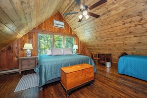 Ein gemütliches Schlafzimmer im Dachgeschoss, mit schrägen Wänden, einem Bett mit blauer Decke, einer Holzablage und einem Schreibtisch mit Stuhl.