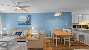 A modern living room with a sofa, a dining table, and a kitchen area with cabinets and a microwave. - Poolside Condo with Beach Access (Gulf Shores)
